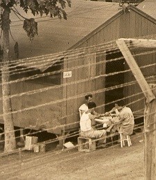 sepia photo of three people sitting at a table outside a wooden building behind a tall barbed wire fence