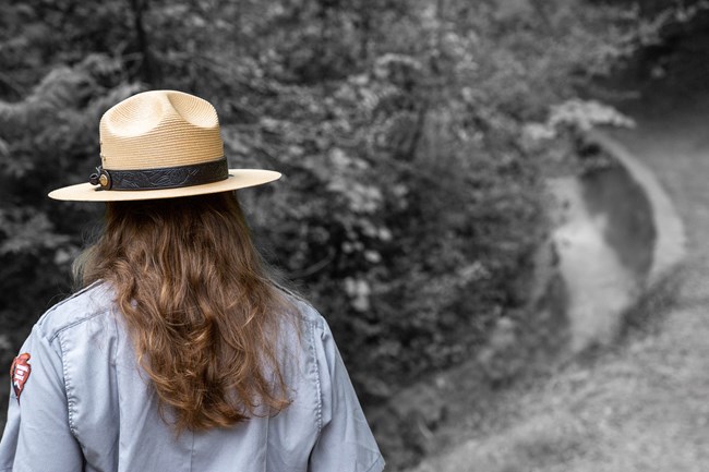 A woman with long brown hair wears a tan straw flat hat as she looks at water in a cascading culvert.