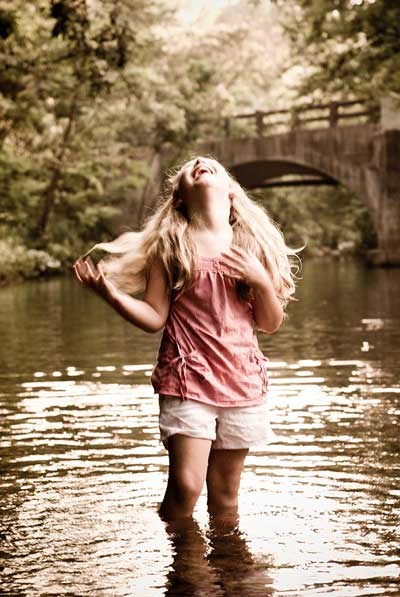 Tinted photo of a young girl with long blond hair standing in a stream; her head is back in laughter. Behind her is a bridge and treeslining the stream. The girl is wearing a pink top and white shorts.