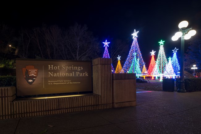 Sign that reads Hot Springs National Park with decorated trees to the right at nighttime