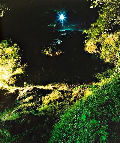 photograph of the hot spring cascade at night, making it a dark photo. There is green vegetation showing on three sides around the dark water and at the top, where there is a black void, there is a white star shape.
