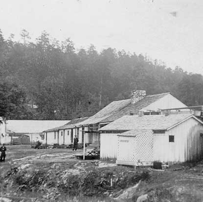 black and white photo of small white wooden buildings buildings built on the right side of the creek bank.