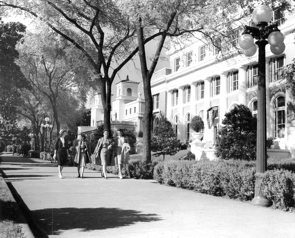 Black and white photo from the late 1940s of 4 women walking down the sidewalk of bathhouse row in dresses. They are in front of a bathhouse with 4 pillars, bushes and trees. Trees line both sides of the walk.