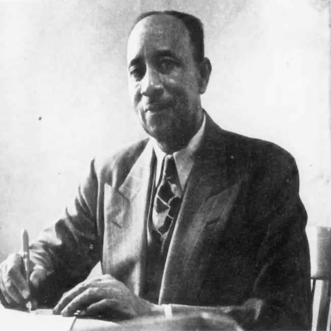 Balding man sits at desk holding a pen and wearing a white shirt, dark tie, and dark suit.