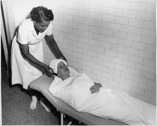 African American woman wearing a white uniform applies towel to a white woman's head who is laying on a bed wrapped in a white towel.