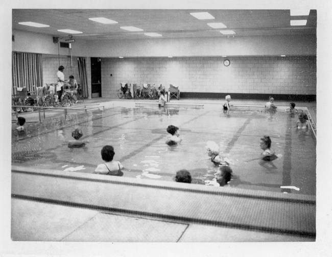 Thirteen people stand in a shallow pool. A nurse helps a man in a wheelchair outside the pool.