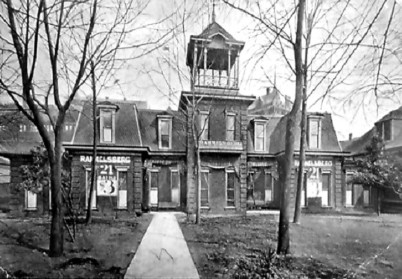 Old wooden, two-story Rammelsberg Bathhouse circa 1900 with multiple trees on lawn in front of building. Sign on building advertises 21 baths for $3.