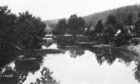 Historic black and white photo of the lake at whittington park in 1897. It is lined with trees reflected in the water and there is a white pagoda on the far side.