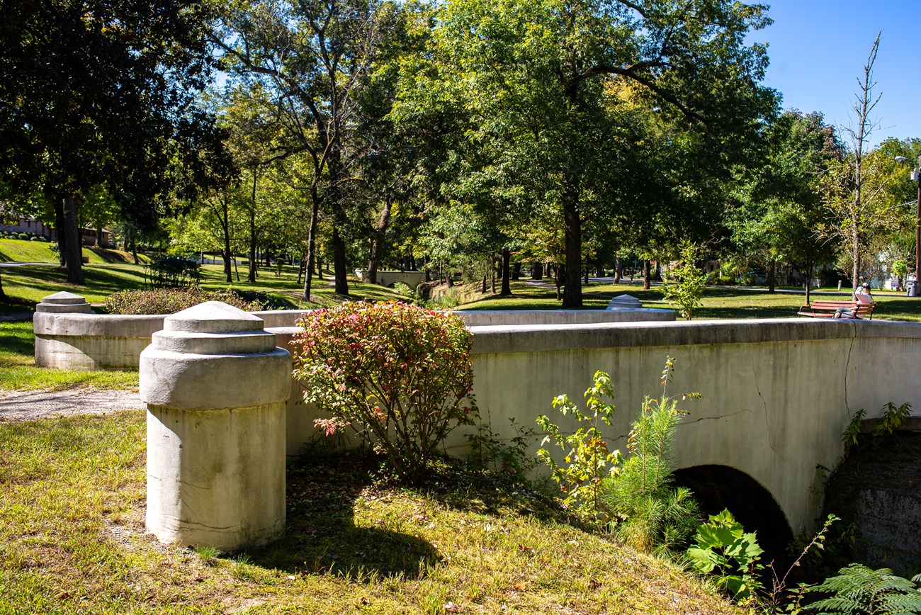 White stone bridge, with 2 pillars at the entrance, arches over Whittington creek. Green plants grow up the hill from the creek bed. Tall trees with green leaves stand in the distance outlined by blue sky.