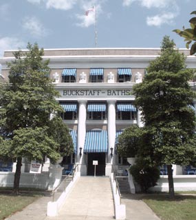 color photo of Buckstaff, showing the ramp and center front of the building. It is a three story building of grayish-brown brick and white Doric colums with classic urns flanking the top of the ramp and between the third floor windows. It has blue and white striped awnings. color photo of Buckstaff, showing the ramp and center front of the building. It is a three story building of grayish-brown brick and white Doric colums with classic urns flanking the top of the ramp and between the third floor windows. It has blue and white striped awnings.