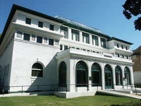 color photo of the Maurice from the north front side. It is a three story white stucco building of the California Modern style with large arched plate glass windows on all sides of the enclosed porch. Center of the roof you can see the skylight cover peaking over the top of the green tiled roof.