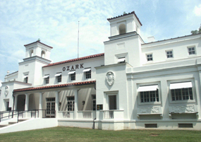 color photo of Ozark Bathhouse from the south end of the building, with the sidewalk and lawn in front. It's a sunny day with a light blue sky with a few clouds. The Ozark is a white stucco building of the Spanish Colonial Revival style of architecture with a red tile roof and open porch. color photo of Ozark Bathhouse from the south end of the building, with the sidewalk and lawn in front. It's a sunny day with a light blue sky with a few clouds. The Ozark is a white stucco building of the Spanish Colonial Revival style of architecture with a red tile roof and open porch.