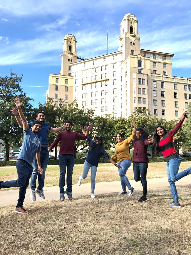 A group of 8 friends jump on a lawn. A tall, looming hotel is in the background.
