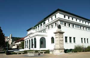 color photo of Maurice Bathhouse from southwest corner of building showing fountain and entrance pylon at historic formal entrance. The Maurice is a three story white stucco building with a skylight frame showing on front center side of roof and green roof tiles.