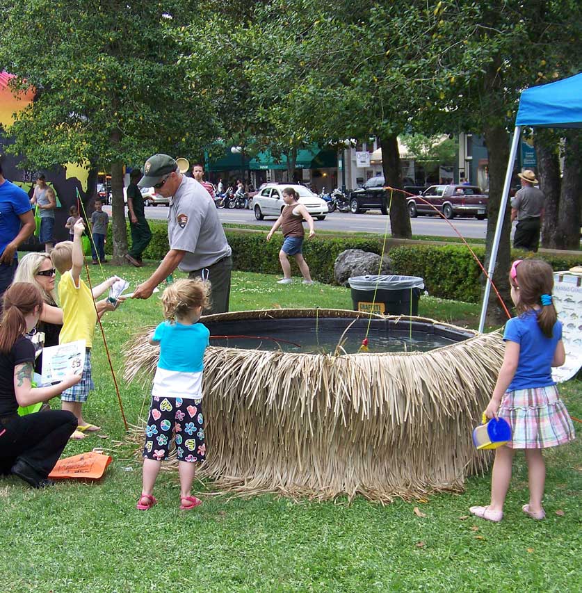 Children standing around a large round tub that has a grass skirt. Ranger is helping child with fishing pole.