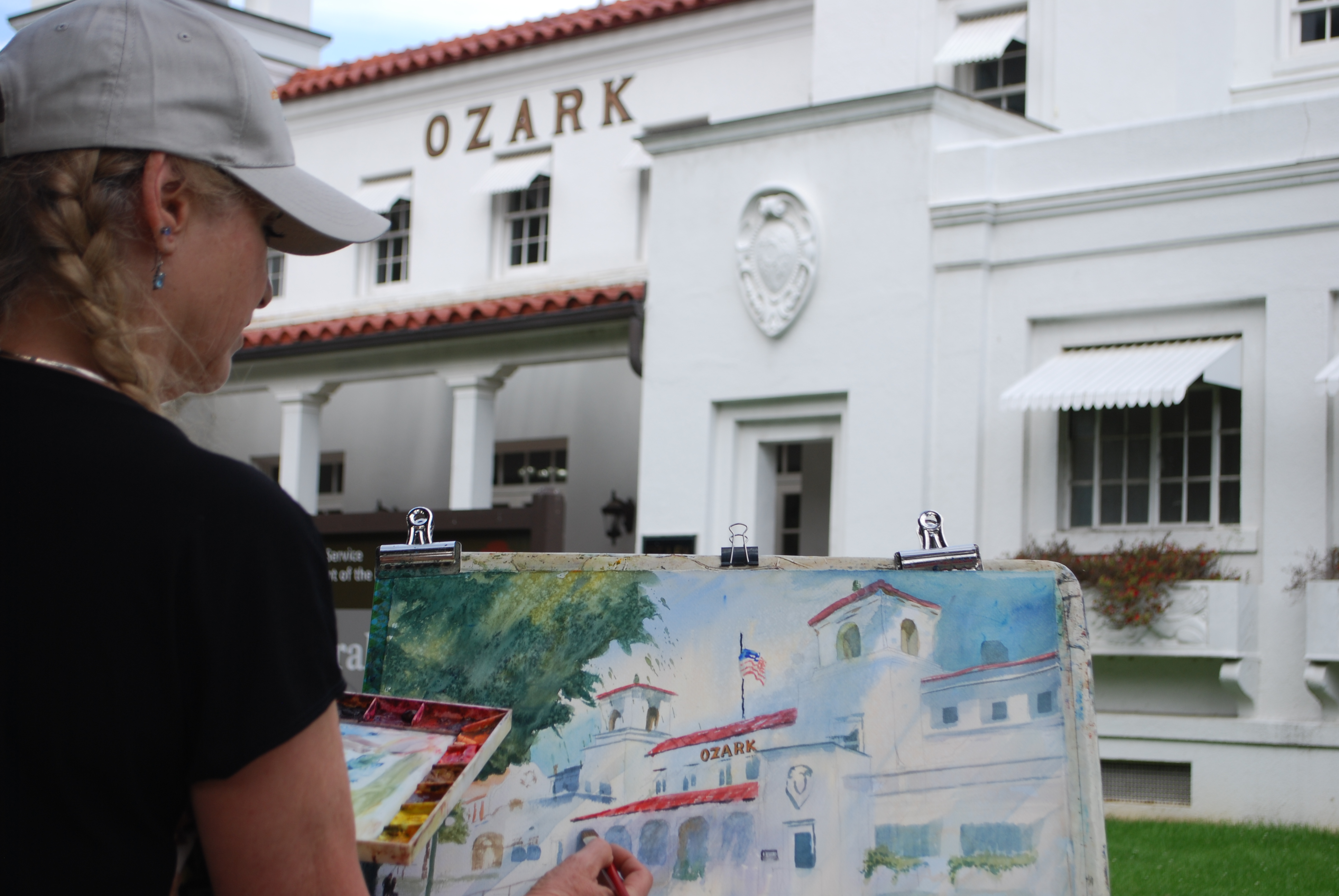 A woman paints a white bathhouse on an easel while standing outside that bathhouse.