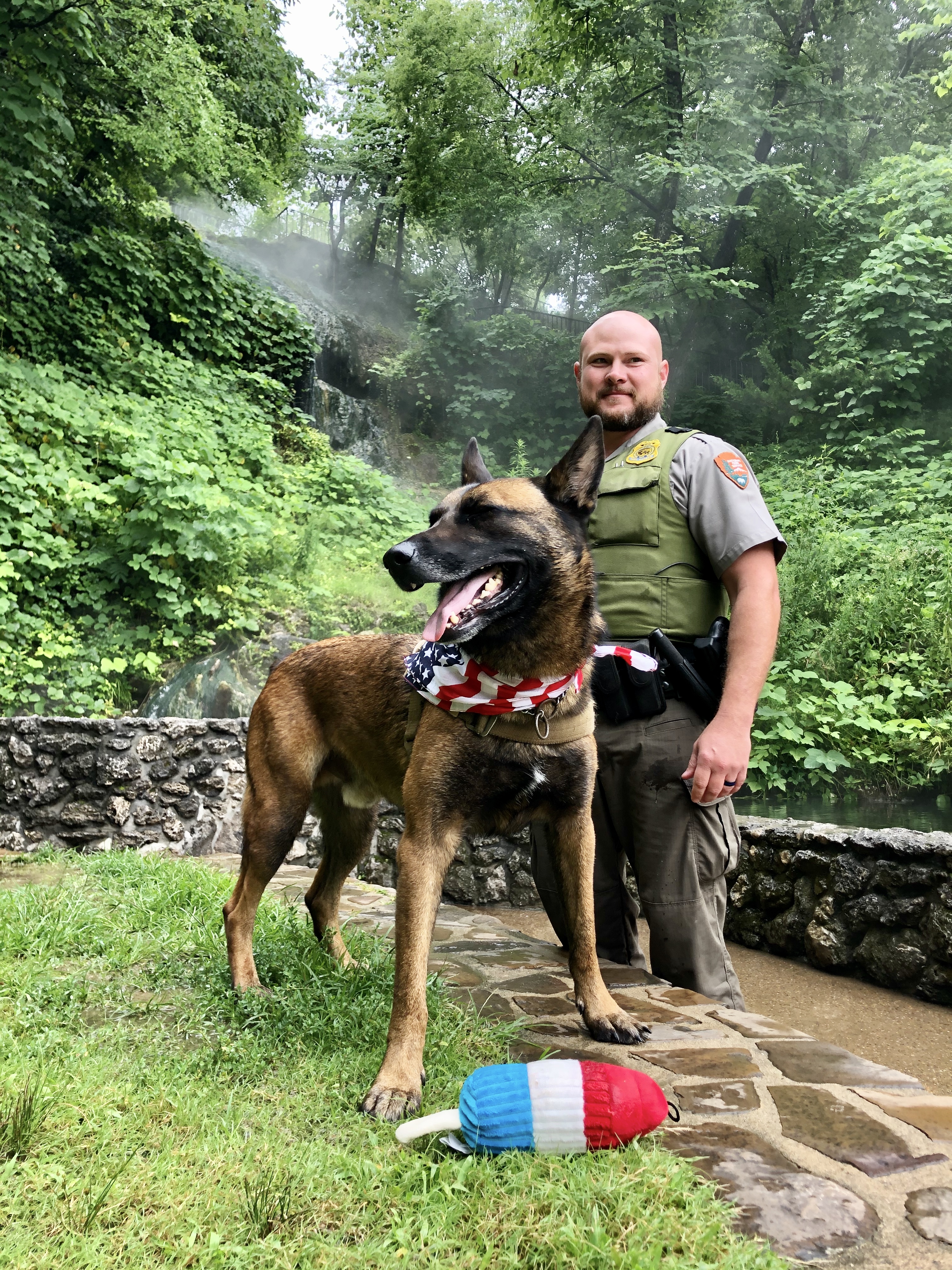 A dog wearing a red, white, and blue bandana stands next to a man in a ranger uniform.