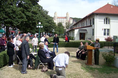 sSall crowd seated and standing on left, park superintendent at lecturn on right. Two women seated behind lecturn. On grassy lawn with white building in background.