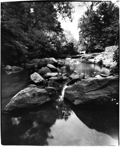 Black and white photo (taken with a pinhole camera so the edges are not square) of a Gulpha Creek looking upstream from a pool. There are large rocks flanking the creek with a small fall near the center. The artist is in the center but blurry so hard to see. Trees close in in the background.