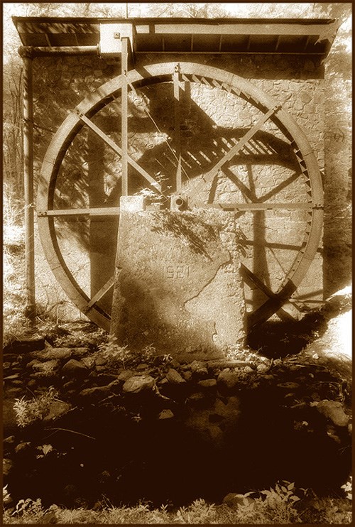 sepia tone photo of a stone structure with a large water wheel mounted on the side, shadowy foreground