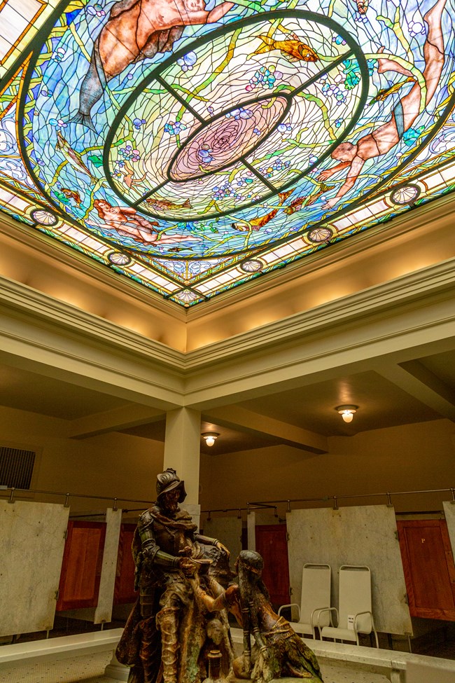 The exquisite stained glass ceiling of mermaides swimming in a pond over a statue of a native american woman offering water to a traveling man