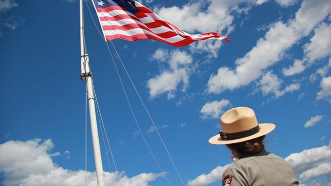 A female park ranger wearing a straw stetson hat looks up at the american flag, red white and blue, waving in the wind against a bright blue sky.