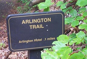 Close up view of Arlington Trail sign, a brown metal sign with gold lettering and a gold border line, surrounded on the right by leafy foliage Close up view of Arlington Trail sign, a brown metal sign with gold lettering and a gold border line, surrounded on the right by leafy foliage