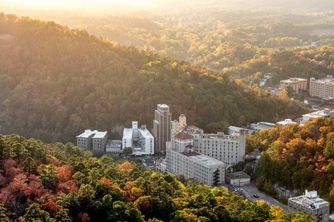 An elevated view looking over Hot Springs with rolling hills, tall evergreen pine trees and blooming deciduous trees. The sun is shining and casting a glow over the valley.
