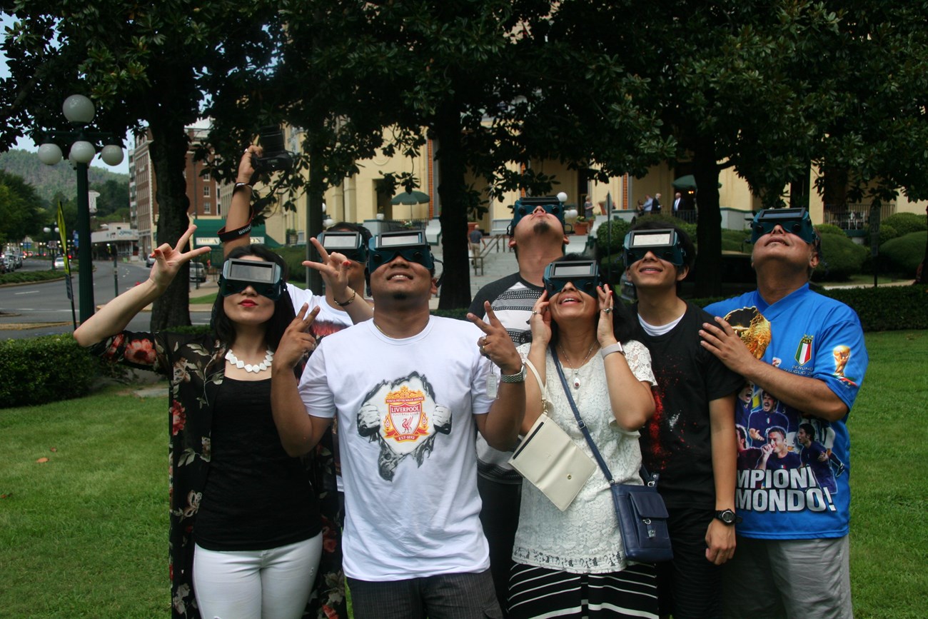 A family of 7 wearing Eclipse goggles happily look up at the sky.