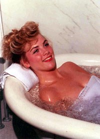 Large ceramic white bath tub, typical of those in the traditional bathhouses, with a large faucet on the left and a grab bar on the gray marble wall behind the tub;blond haired woman in the tub