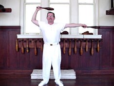 Man standing in old gymnasium in white undershirt and white pants with an Indian club in each hand.