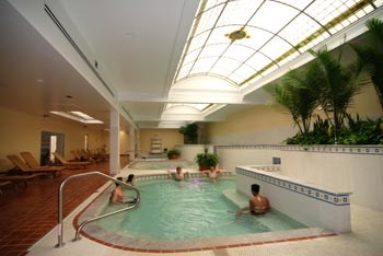 pool area in Quapaw shot from one end. On right is tiled lounging area with wooden cots and on left are several connected pools. overhead on right is a curved stained glass window.