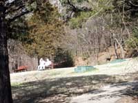 spring scene of grassy hillside with cement trail on lower right and two green boxes sticking up from ground to left of trail, visitors sitting on a wooden park bench in front of a cedar tree on far left, with wooded hillside in background spring scene of grassy hillside with cement trail on lower right and two green boxes sticking up from ground to left of trail, visitors sitting on a wooden park bench in front of a cedar tree on far left, with wooded hillside in background