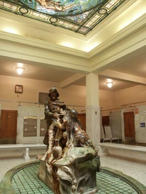 color photo of Men's Bathhall in Fordyce Bathhouse, with bronze colored ceramic fountain depicting a conquistador with an American Indian in the foreground and a portion of the stained glass skylight ceiling. color photo of Men's Bathhall in Fordyce Bathhouse, with bronze colored ceramic fountain depicting a conquistador with an American Indian in the foreground and a portion of the stained glass skylight ceiling.