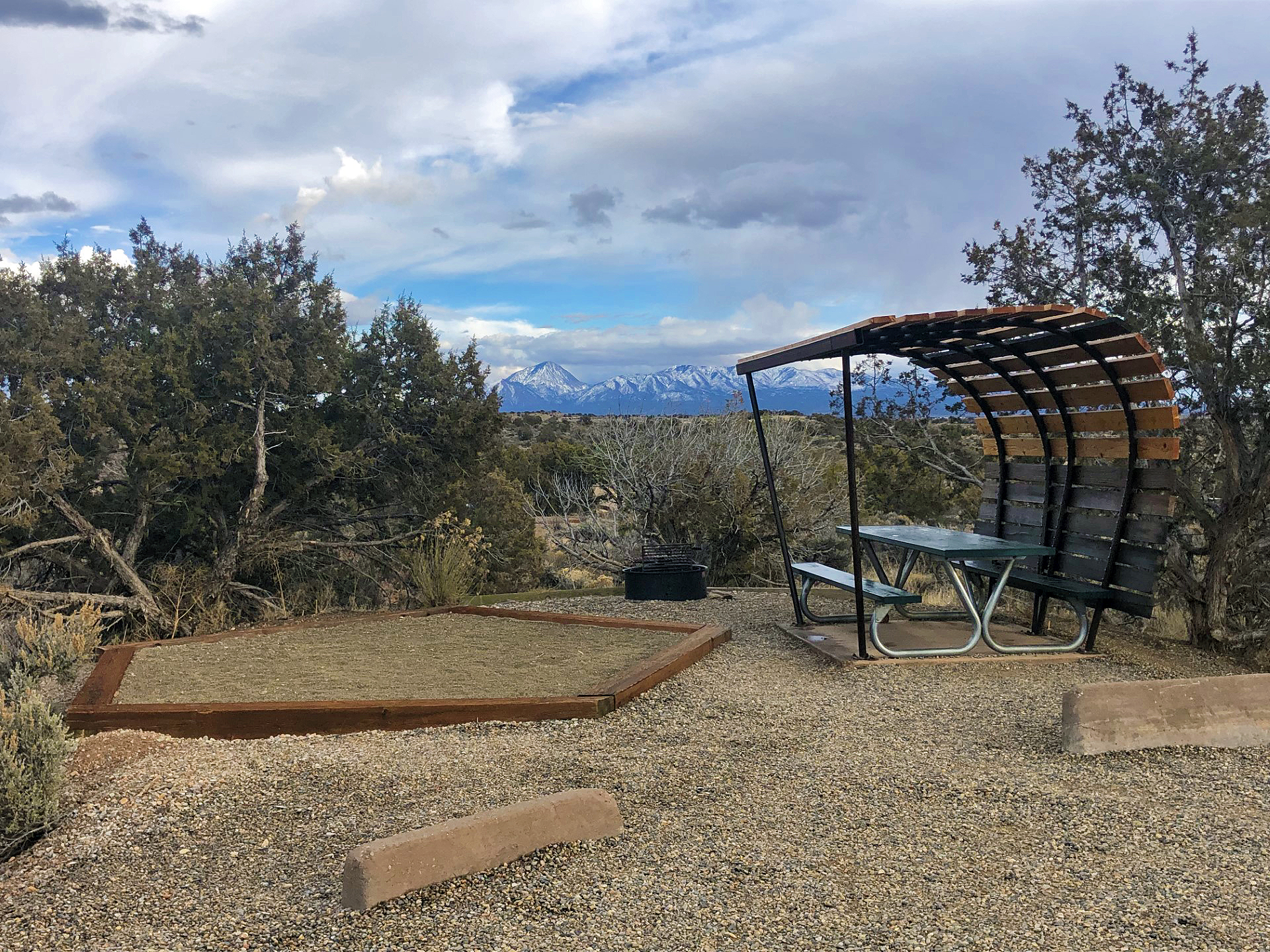 a gravel campsite with tent pad, shade-covered picnic table, and juniper tree; mountains in background