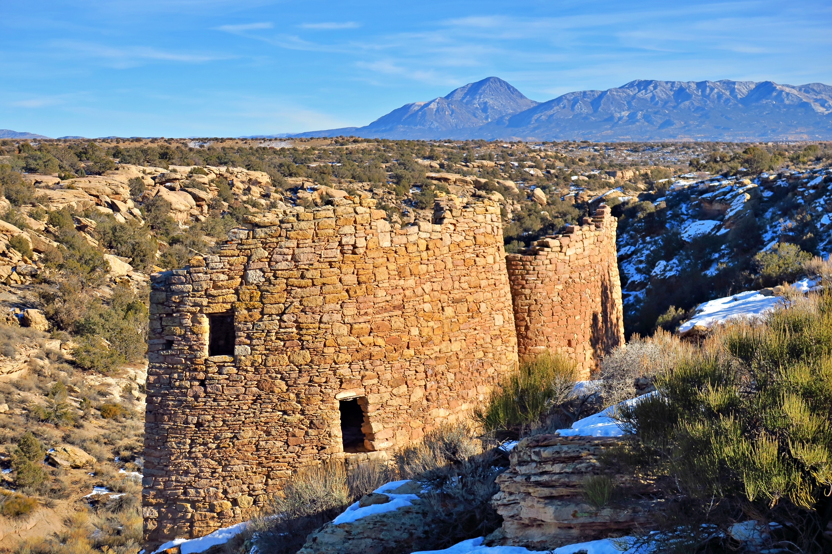 Winter scene with an ancestral puebloan structure, patchy snow around it, and mountains in the background