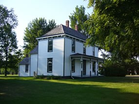 Truman Farm home in a modern photograph, surrounded with green grass and full trees. The Home itself is white, two story, with green trim.