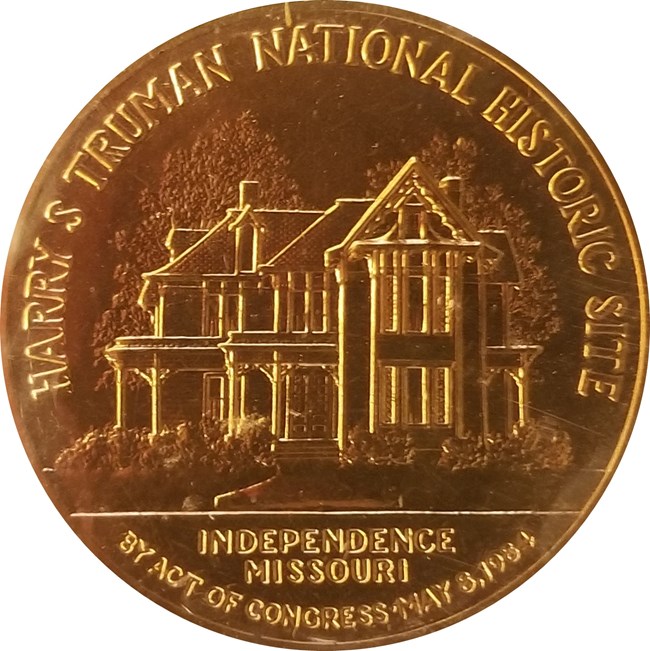 A gold colored coin has an image of the Truman Home, with park name and Act of Congress May 8 1984  Independence Missouri