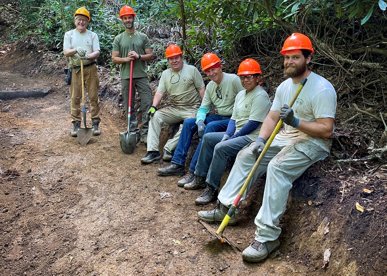 Six people smiling along a trail, each wearing a hard hat, some holding shovels and trail maintenance equipment. Green rhododendron and other vegetation line the trail.