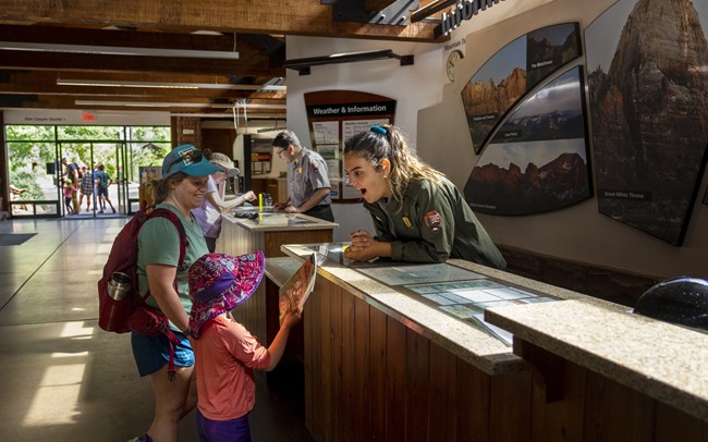 Photograph of NPS ranger behind desk smiling at child and mother with Junior Ranger booklet.