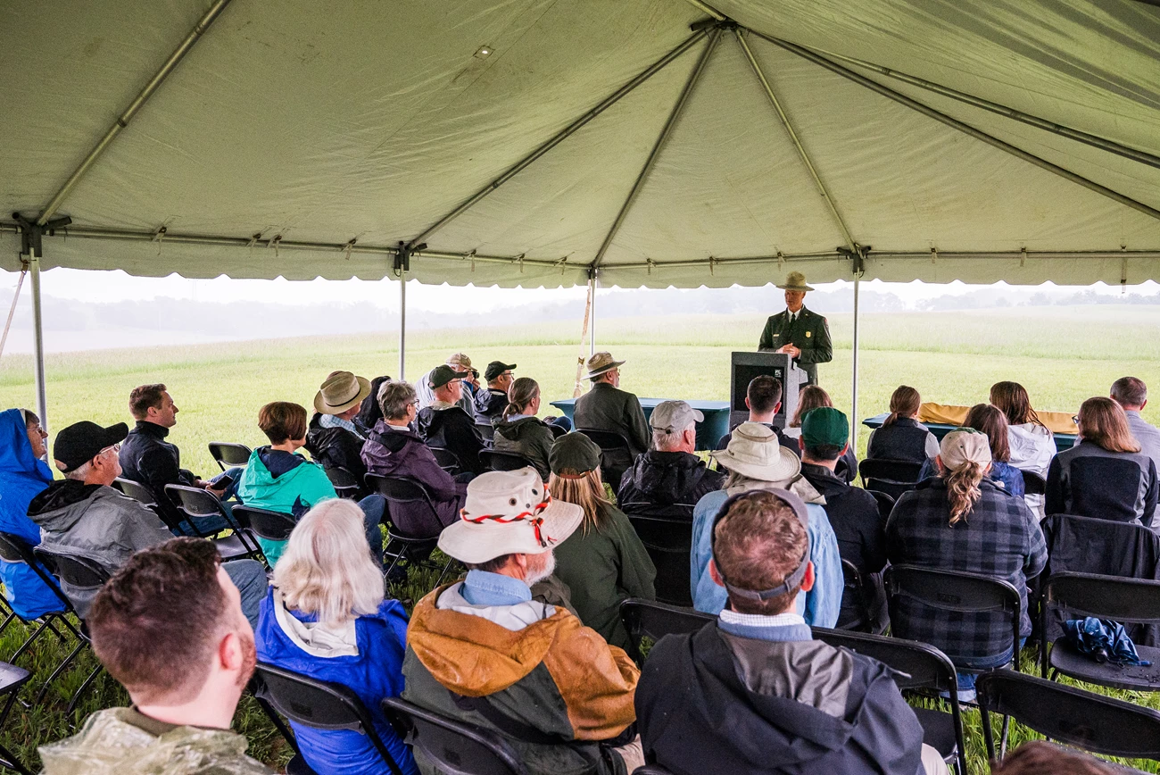 Photograph of a person in a National Park Service uniform stands at a podium under a tent in front of a group of seated people. The background shows an outdoor rainy scene.