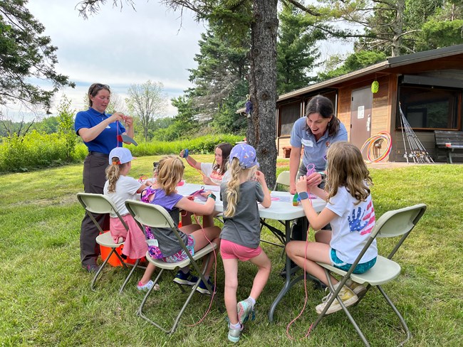 On a lawn, next to a tree, with a building in the background, two women stand beside a table with young girls seated around it as they all work on a project together.