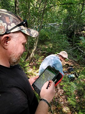 2023-Sept_Hester-Parr_Tablet Data Form_MODIFIED A man in a blue shirt and wide-brimmed hat looks at a green box of equipment while kneeling on the ground. A man in a black t-shirt and ball cap stands behind him entering information into a tablet.