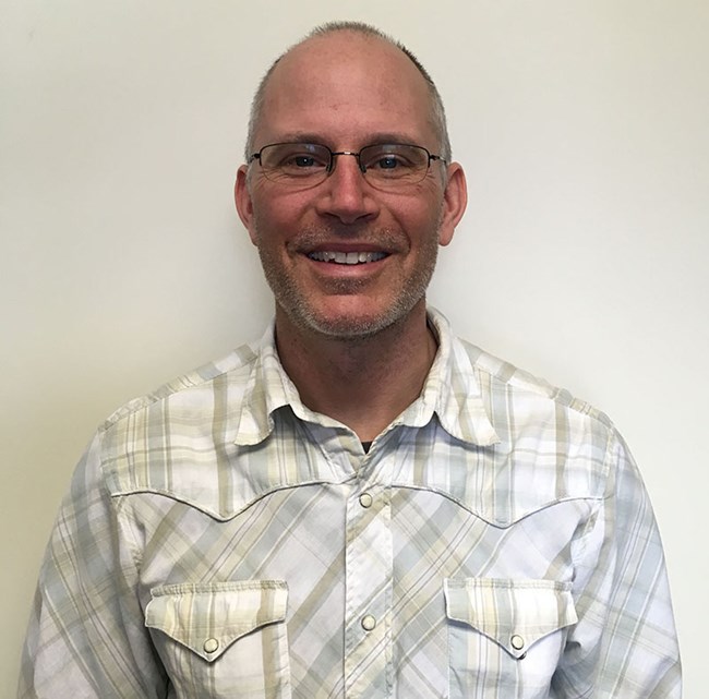 a smiling man stands against a blank wall