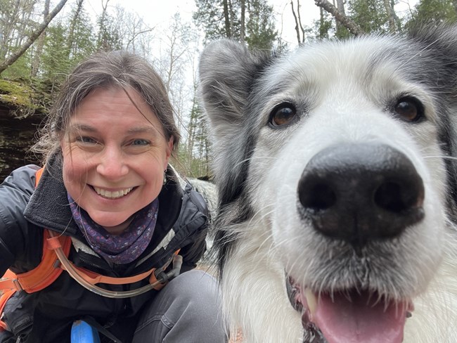 A smiling woman sits beside a gray and white dog whose face takes up the right half of the photo. Conifer trees are in the background.