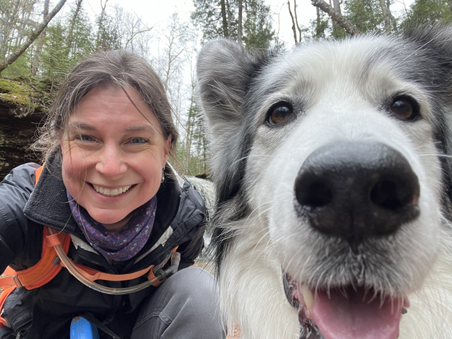 A smiling woman sits outdoors next to a white and gray dog.