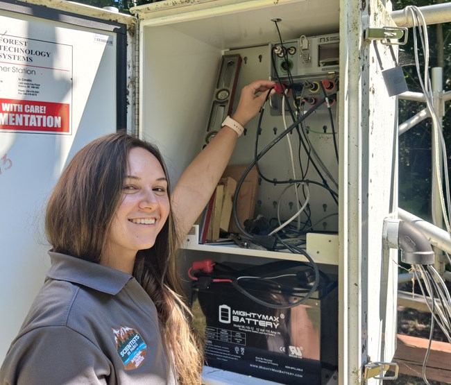 A woman with long brown hair smiles as she plugs sensors into a control panel at the base of a weather station.