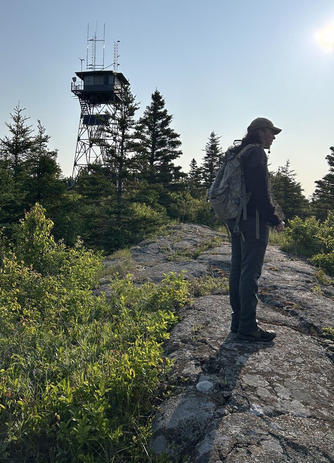 A man wearing a daypack stands on a rocky outcrop in morning sunlight. A lookout tower stands at the top of the hill beyond him.