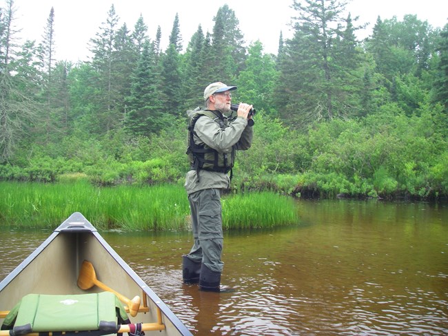 A man stands in a river beside a canoe holding binoculars.