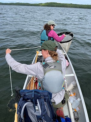 Two women in a canoe pull up nets attached to a tow line. Trees line the distant shoreline.